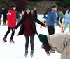 A group of people is enjoying their time on an ice skating rink with a smiling woman in the foreground showing off her balance while another person appears to be bending down or falling in the foreground