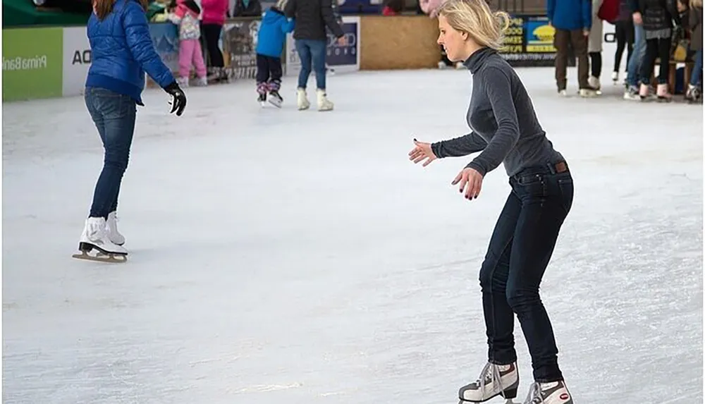 A woman in a gray top and jeans is ice skating at an outdoor rink appearing focused as she glides on the ice