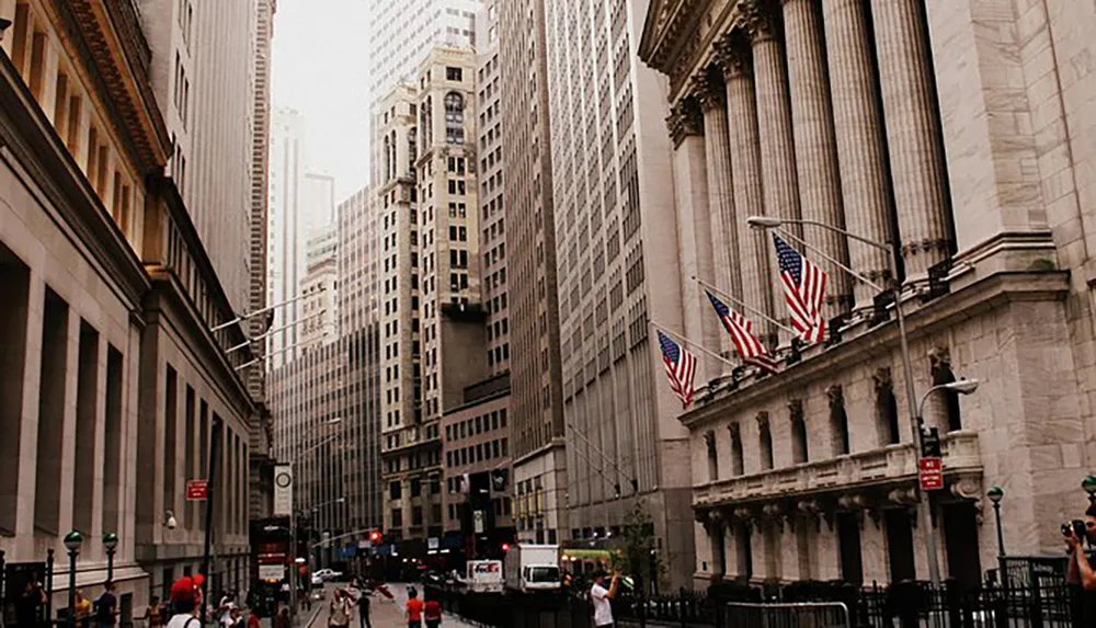 This image shows a bustling urban street flanked by towering buildings with American flags prominently displayed on one of the classical facades suggesting a location of significance possibly in a financial district