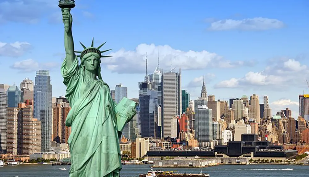 The Statue of Liberty stands in the foreground with New York Citys skyline as the backdrop under a clear blue sky