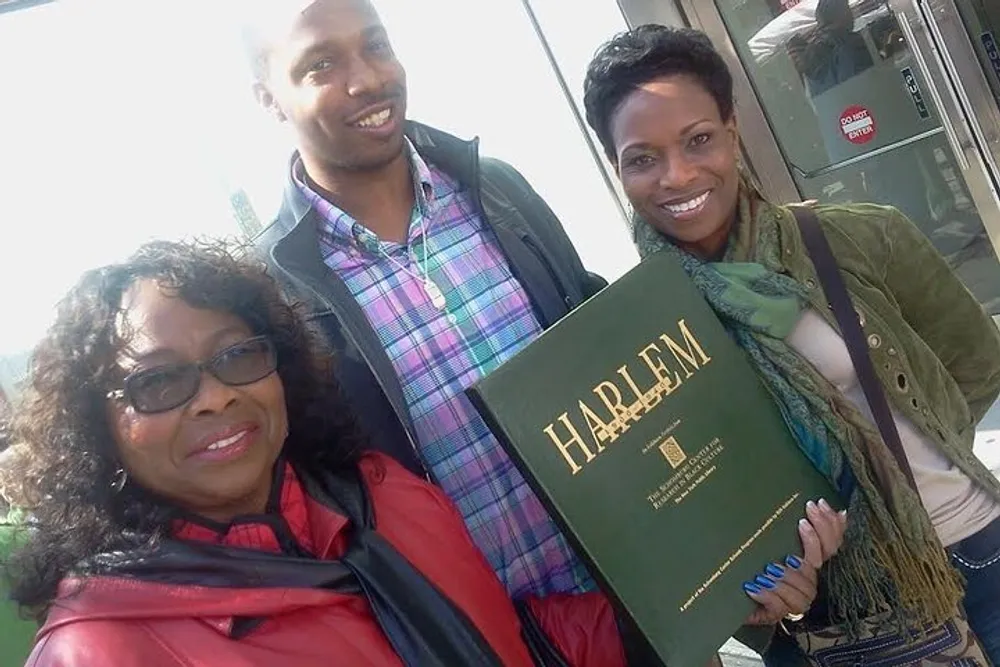 Three smiling people are posing with a book titled HARLEM with a woman in the center holding the book