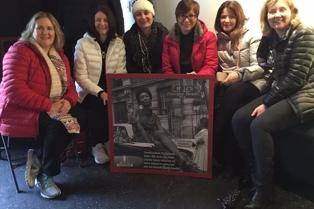 A group of six women are smiling and posing for a photo with one of them holding up a framed black and white street photograph with a placard