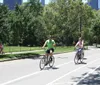 People are enjoying a sunny day bicycling in a park with trees and a clear blue sky with a glimpse of tall buildings in the background