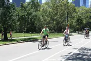 People are enjoying a sunny day bicycling in a park with trees and a clear blue sky, with a glimpse of tall buildings in the background.