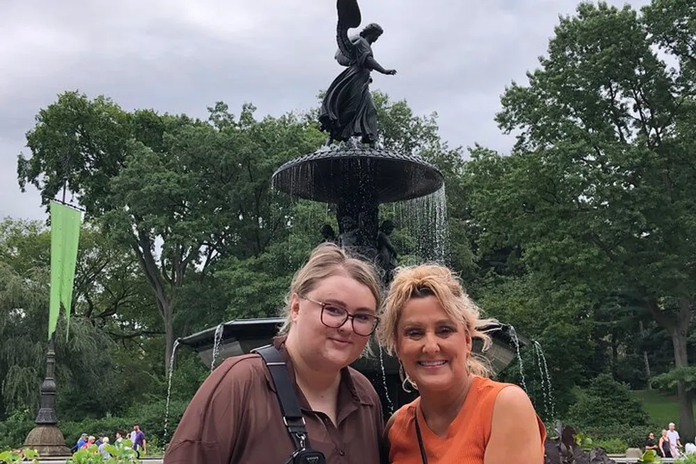Two women are smiling for a photo in front of a fountain with a statue on it in a park setting