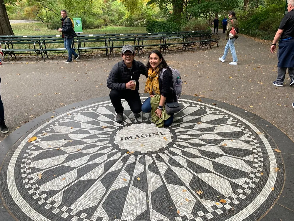 Two people are smiling and posing for a photo on the Imagine mosaic memorial in Central Park