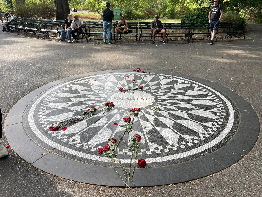 The image shows a mosaic with the word IMAGINE at its center adorned with scattered red roses surrounded by people sitting and standing around it in what appears to be a park