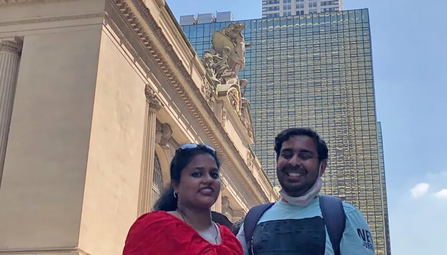 Two people are smiling for a photo in front of a classical building facade with a modern skyscraper visible in the background.