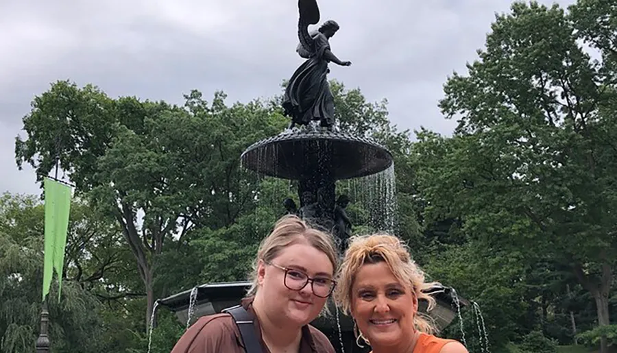 Two smiling individuals are posing for a photo in front of a water fountain adorned with a statue, surrounded by lush greenery.