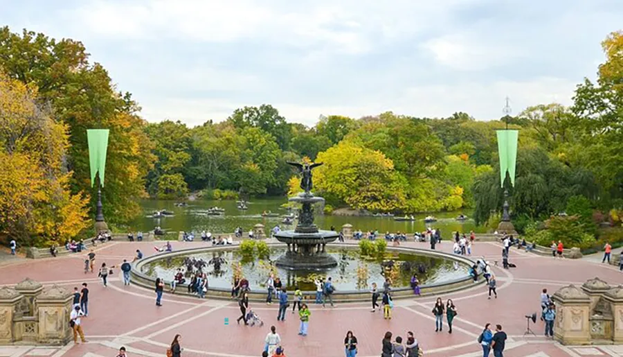 The image captures a bustling scene around the Bethesda Fountain in Central Park, New York City, framed by autumn foliage and a clear sky.