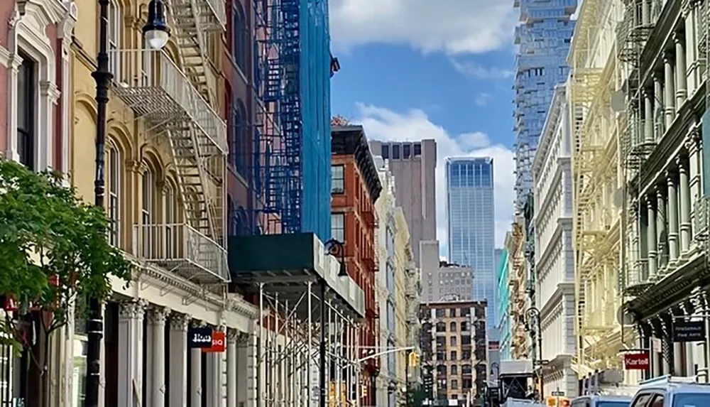 The image captures a vibrant urban street lined with historic buildings featuring fire escapes with the contrast of modern skyscrapers in the background under a clear blue sky