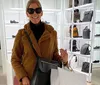 A person is smiling for the camera inside a boutique with bags and shoes on display wearing sunglasses and a teddy coat and holding a shopping bag