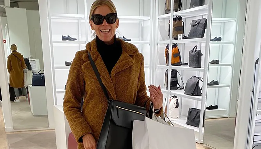 A person is smiling for the camera inside a boutique with bags and shoes on display wearing sunglasses and a teddy coat and holding a shopping bag