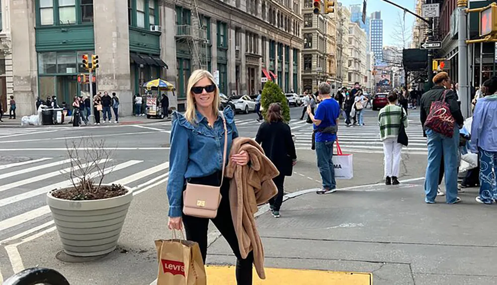 A smiling person stands on a city street corner with shopping bags with other pedestrians and an urban backdrop around them