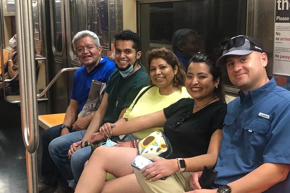 Five individuals are smiling and sitting side by side inside a subway train appearing to enjoy one anothers company