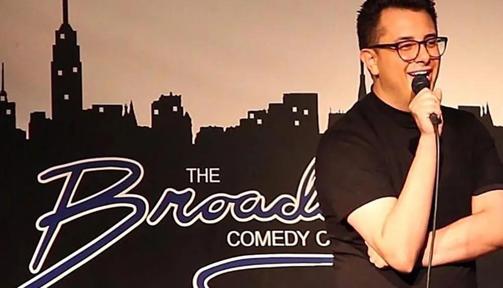 A person is performing with a microphone at The Broadway Comedy Club with a backdrop featuring a city skyline silhouette