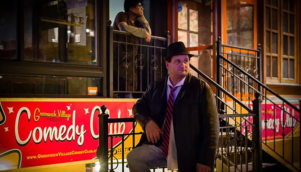 A man wearing a jacket and a hat is sitting on the steps outside the Greenwich Village Comedy Club during the evening