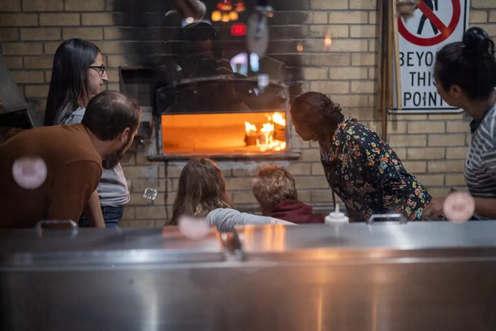 Customers are attentively watching a chef prepare food in a wood-fired oven at a cozy restaurant