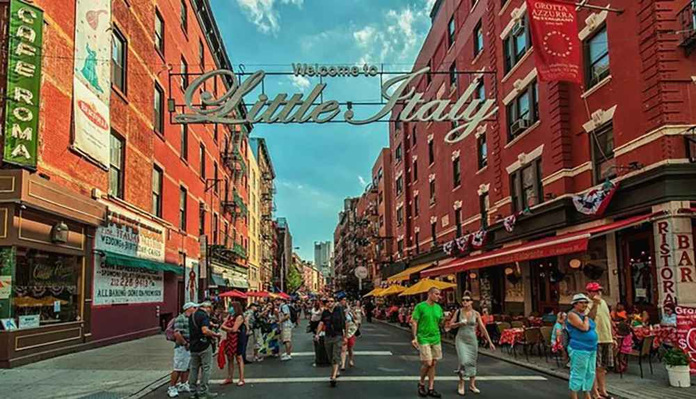 The image shows a vibrant street scene from Little Italy with people outdoor dining and a Welcome to Little Italy sign overhead