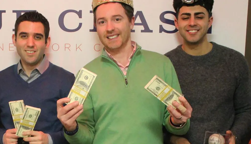 Three men are smiling for the camera while holding stacks of money with the central figure wearing a playful crown