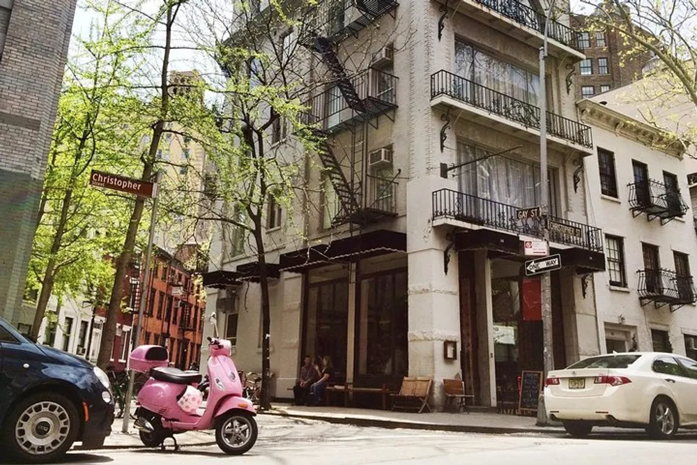 This image shows a sunny street corner with a pink scooter parked in front of a building with a cafe at street level pedestrians sitting outside and street signs indicating Christopher and Gay St
