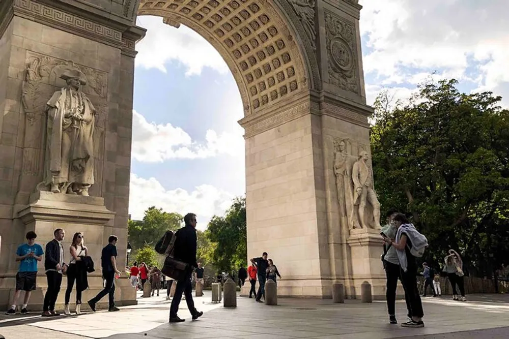 The image shows people passing by and congregating near a large ornate archway on a sunny day likely in a city park