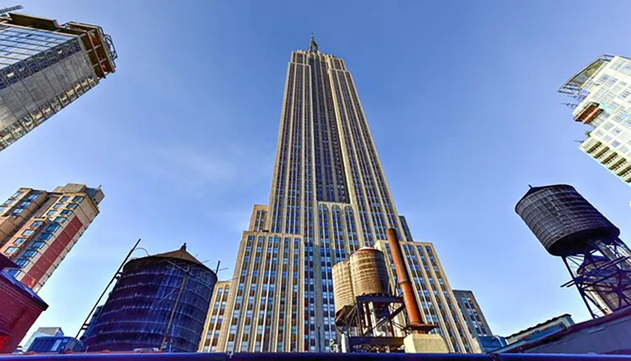 The image shows a towering skyscraper reaching into a clear blue sky, flanked by high-rise buildings and framed by two rooftop water towers.