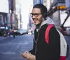 A happy man wearing headphones and glasses is holding a smartphone on a bustling city street