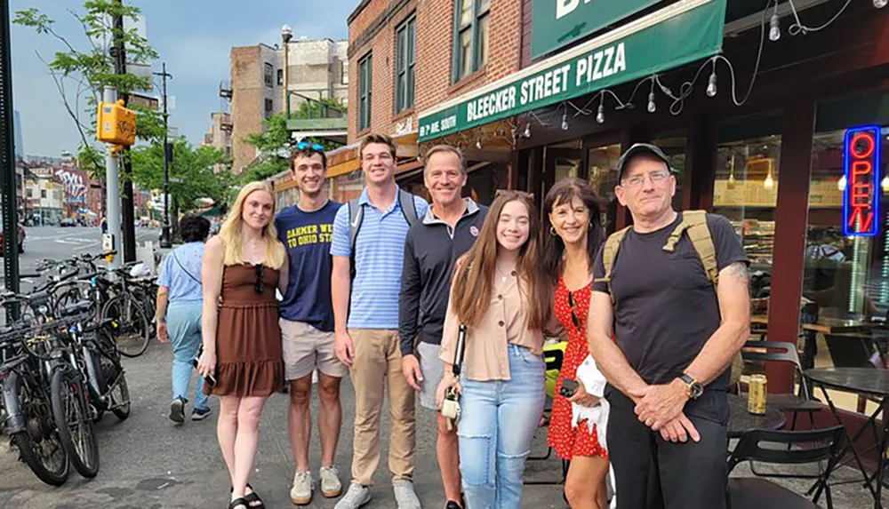 A group of seven people is smiling for a photo in front of Bleecker Street Pizza with bikes and an urban street in the background