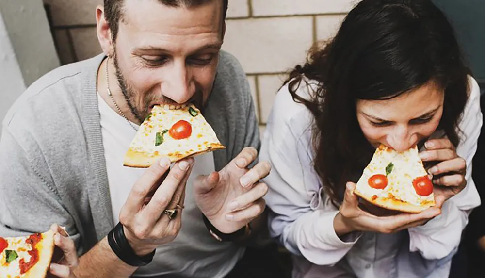 A man and a woman are happily taking big bites out of slices of pizza