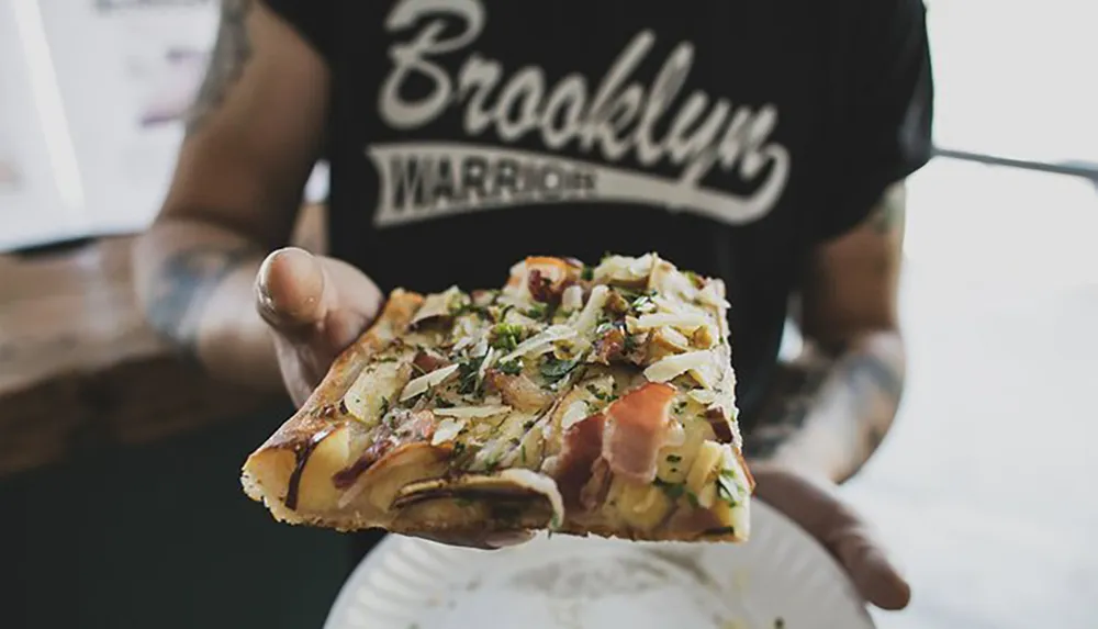 A person wearing a black t-shirt with the text Brooklyn Warriors is holding a slice of pizza with various toppings