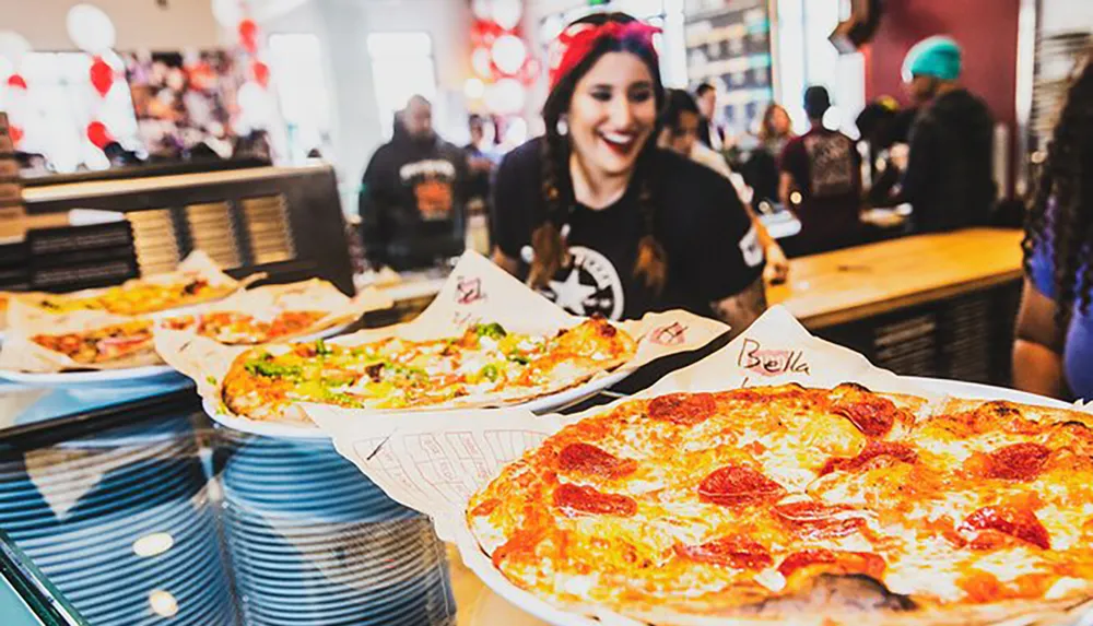 A smiling person with braided hair is in a bustling pizzeria with several large pizzas displayed on the counter in front