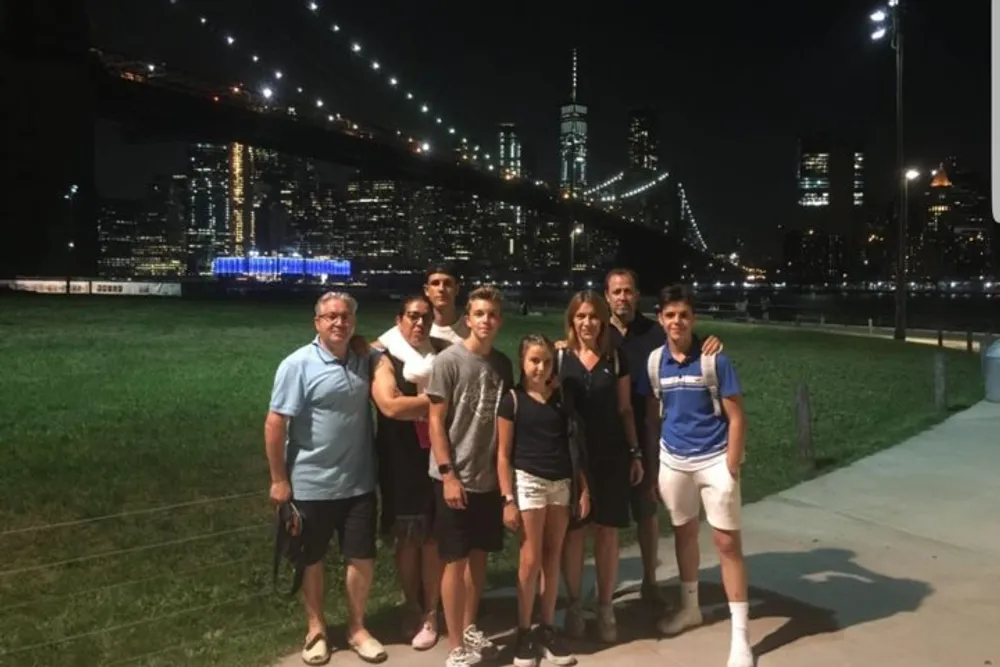 A group of people is posing for a photo at night with the illuminated Brooklyn Bridge and Manhattan skyline in the background
