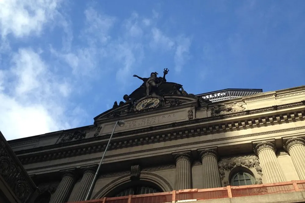 The image shows the upper facade of a grand classical building with the inscription Grand Central Terminal beneath an ornate sculpture and clock framed against a partly cloudy sky