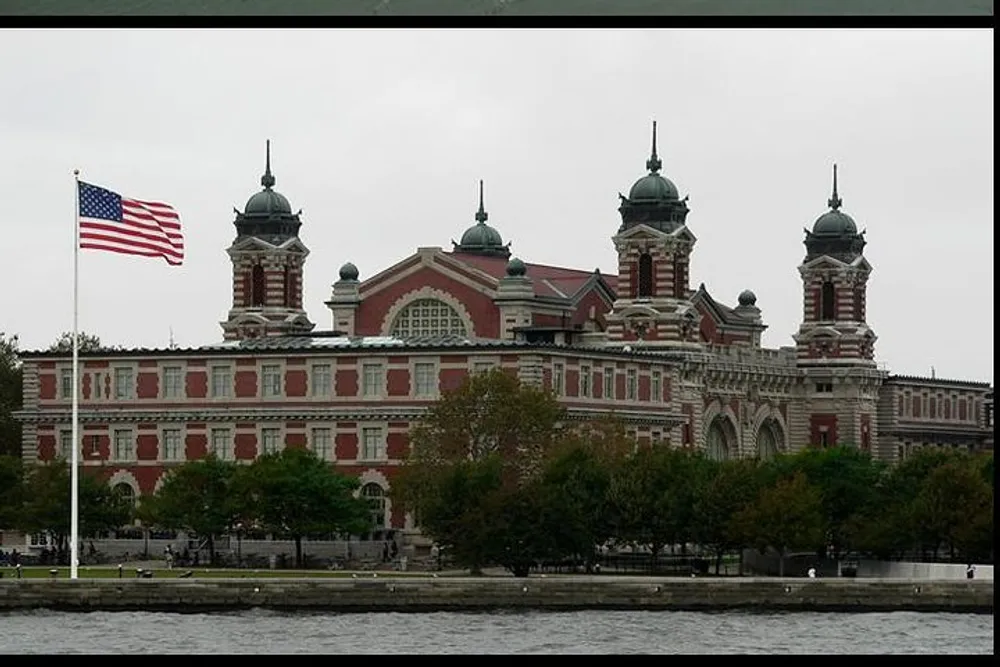 The image shows a large historical building with distinctive red brick and green-topped towers beside a waterfront with an American flag flying prominently in the foreground