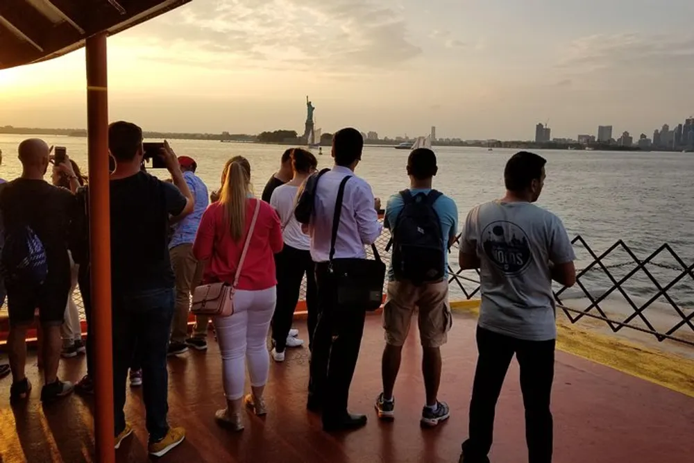 A group of passengers on a ferry are observing and taking photos of the Statue of Liberty during a sunset