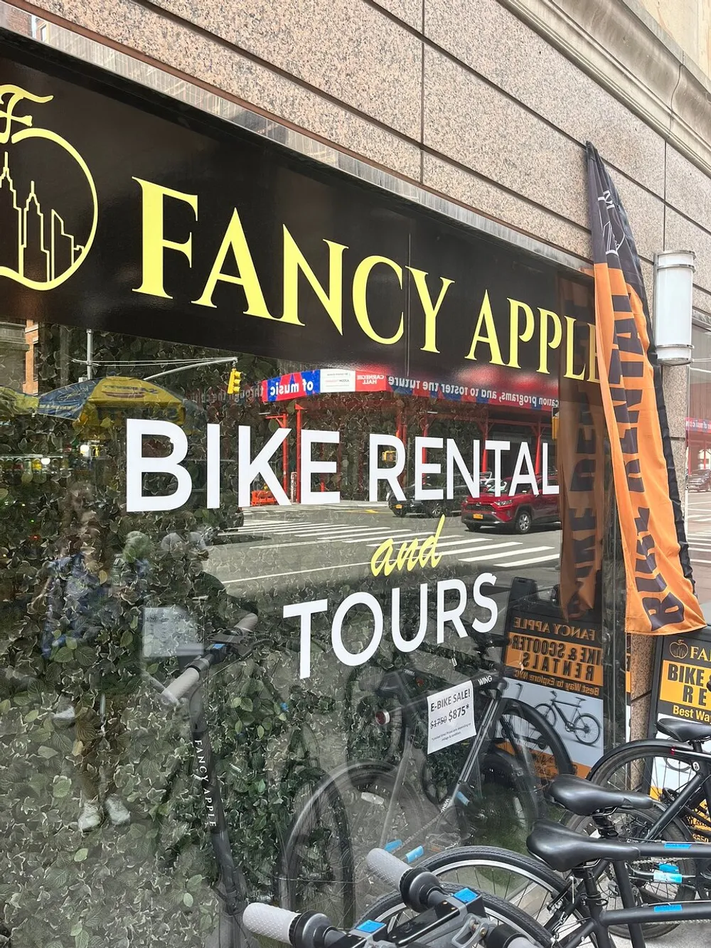 The image shows the storefront of FANCY APPLE BIKE RENTAL and TOURS with bicycles visible through the window and an e-bike sale advertisement reflecting a vibrant urban street scene