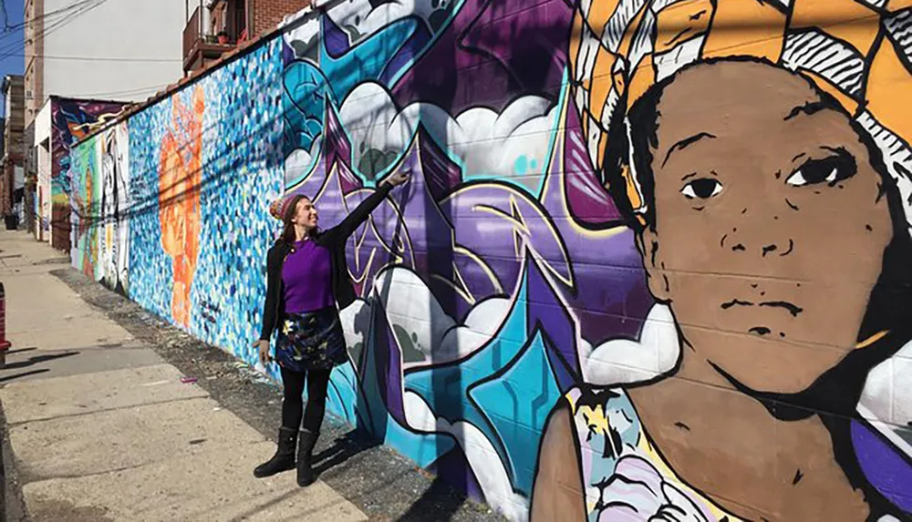 A person is posing in front of a vibrant street mural featuring an expressive portrait and colorful abstract elements