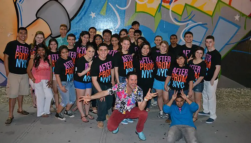A group of cheerful young adults wearing matching AFTER PROM NYC t-shirts poses for a photo in front of a colorful graffiti wall with one person crouching in front displaying a peace hand sign