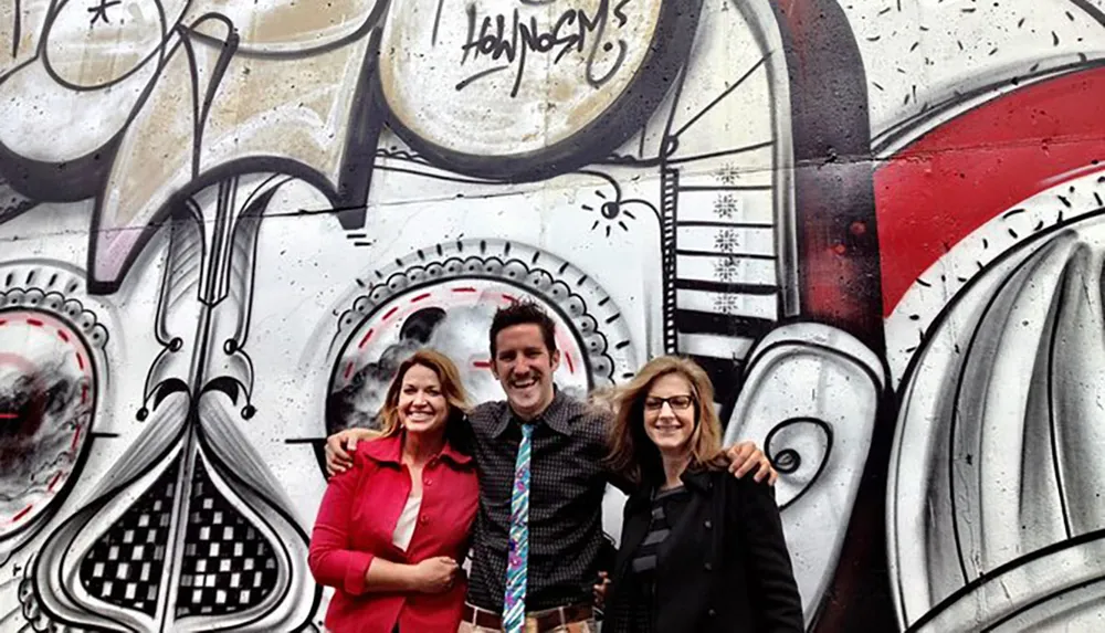 Three people are smiling and posing in front of a wall with large black and white graffiti artwork