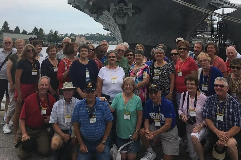 A group of people pose for a photo in front of what appears to be a naval ship with several of them wearing lanyards and casual attire suggesting a group tour or reunion