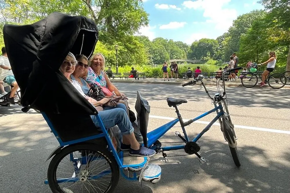 Three people are enjoying a ride in a blue pedicab in a park on a sunny day surrounded by other park-goers and cyclists