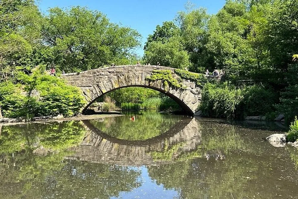 An old stone bridge arches over a tranquil pond surrounded by lush greenery with its reflection shimmering on the waters surface