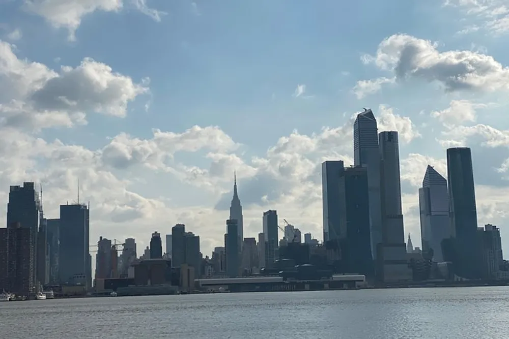 The image shows a daytime silhouette of a city skyline across the water marked by towering skyscrapers and a scattering of fluffy clouds in the sky