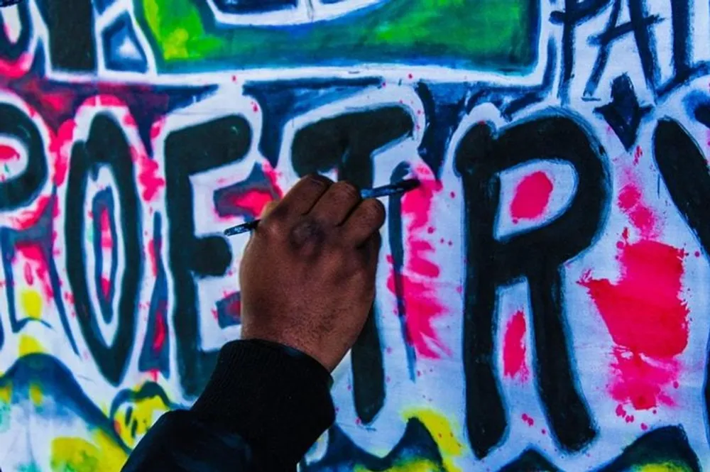 A persons hand is seen painting the word POETRY on a wall covered with colorful graffiti