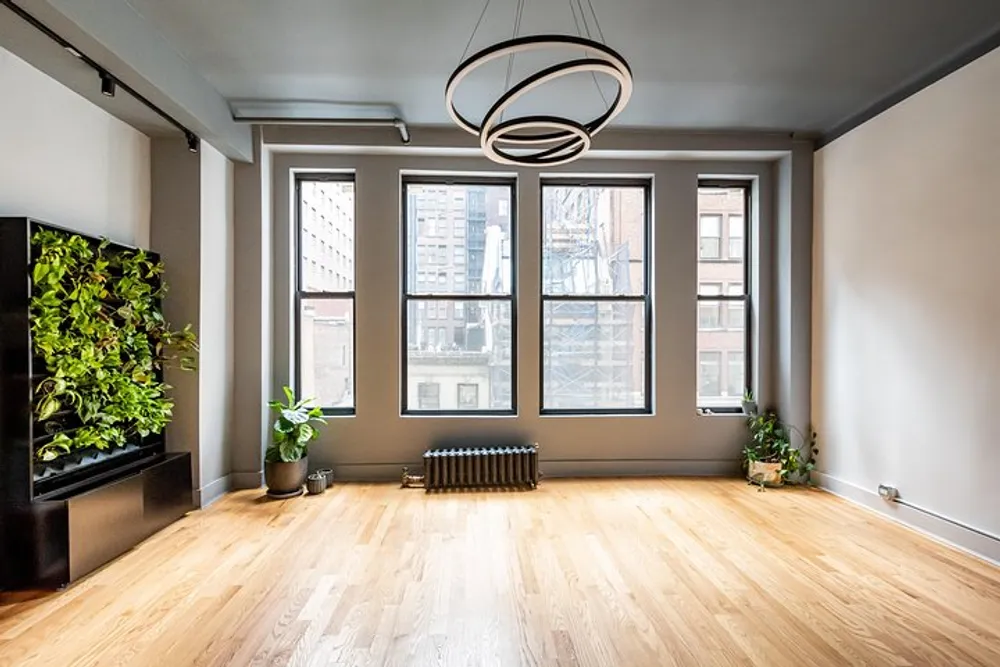 The image shows a modern well-lit empty room with large windows an ornate hanging light fixture and a vertical garden installation on the left wall