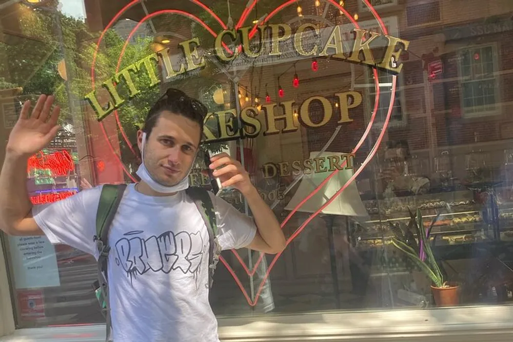 A person is waving at the camera in front of a bakery with a neon sign that reads Little Cupcake Bakeshop