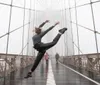 A person is performing a high kick on a foggy bridge with pedestrians in the background
