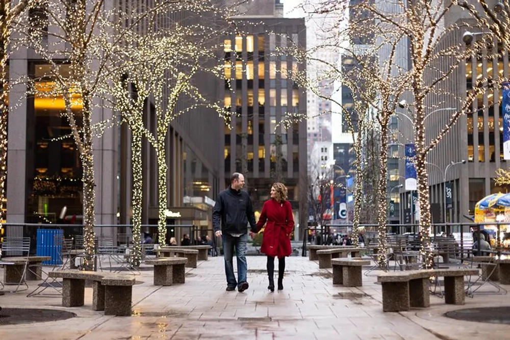 A couple is walking hand in hand through an urban plaza adorned with trees wrapped in twinkling lights creating a romantic setting
