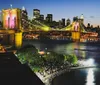 The image shows a vibrant night view of the Brooklyn Bridge illuminated in pink and yellow lights with the Manhattan skyline in the background and a bustling waterfront promenade along the East River